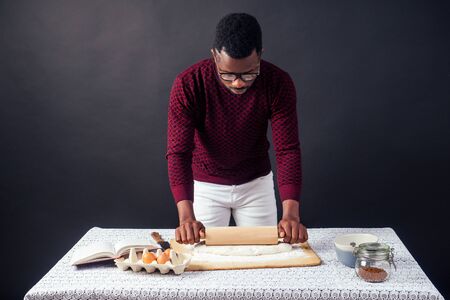 handsome and young afro african man preparing homemade cakes American Pie from fresh dough hands dirty by flour, on the table are eggs, rolling pin and recipe book on a black background in the studioの写真素材