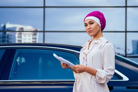 beautiful and successful mulatto afro muslim business woman entrepreneur in stylish hijab and turban holding paper skyscraper office windows background on street standing near her black carの写真素材