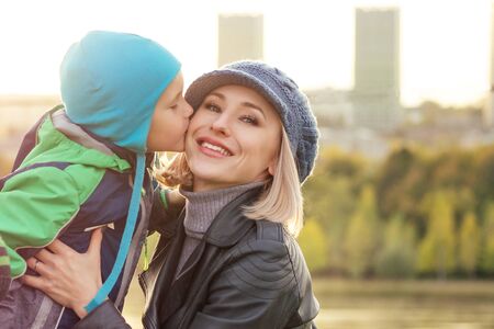 happy and beautiful blonde woman in hat kissing baby son in a warm jacket holding him in her arms in the autumn parkの写真素材