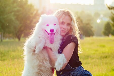 beautiful and charming curly blonde smiling toothy woman in denim shorts are sitting at glass hugging white fluffy cute samoyed dog in the summer park sunset rays field background . pet and mistressの写真素材