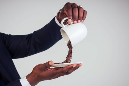 hands of afro american business man in a black classic suit holding a cup with flying coffee bean splash on plate in white background studio shot. Magic morning drinkの写真素材