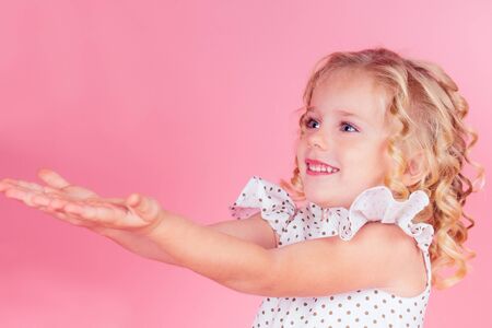 portrait of a beautiful little blonde girl with curly hairstyle and blue eyes in a cute white dress pulling hands on a pink background in the studioの写真素材