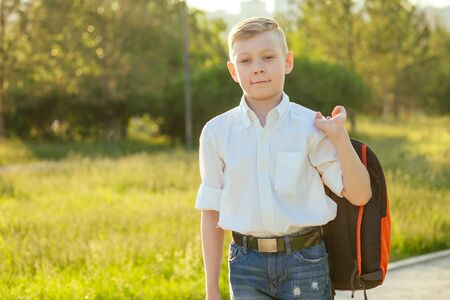 stylish and young schoolboy in a white shirt and jeans with a backpack in the park going to school
