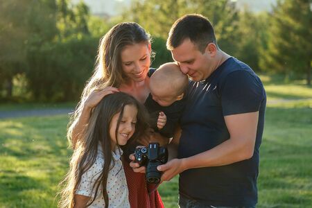 happy family is resting in the park in summer . beautiful mother , handsome father with a little baby boy and cute daughter photographed on camera . family photo shoot watching photos on the screenの写真素材