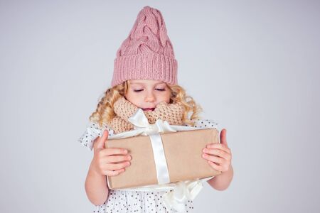 Happy little smiling blonde curly hairstyle girl in a knitted pink hat with christmas gift box with bow on white background in studio. new Year present in the hands of a female child making a wishの写真素材