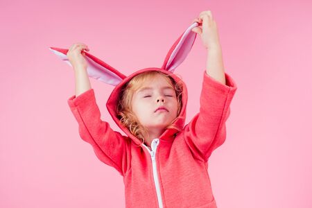 beautiful charming little girl curly blonde hairstyle wearing red sweater clothes with hood rabbit ears.Adorable child celebrate Easter,playing theater role of the easter bunny pink background studioの写真素材