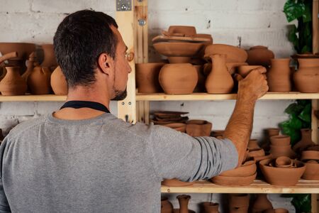 male master potter in apron sculpts a deep bowl of brown clay on a Potters wheel In workshop on the background of shelves with dishesの写真素材