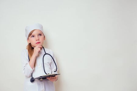 smiling little girl in doctor uniform with medical tools stethoscope writing something to clipboard on white background in studio copy space.future profession vocational guidance careerの写真素材