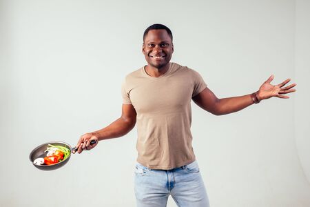 african american chef kitchener holding a frying pan wizard man cooking magic flying food salad, carrot, garlic, onion, pepper, potato, cucumber isolate white background studio.magic of taste fantasyの写真素材