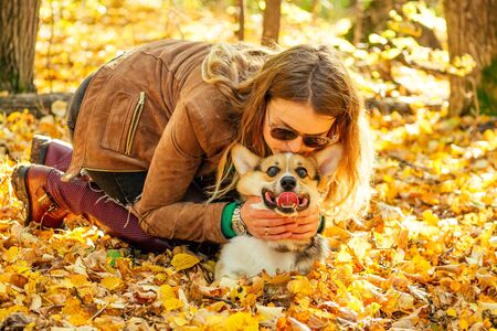 beautiful and young woman in a brown jacket kisses and stylish red boots her pet Corgi in the autumn park . love to the animalsの写真素材