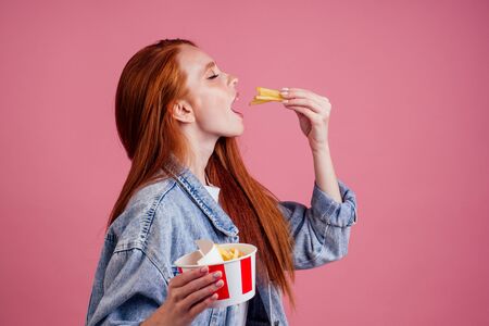 cute red long foxy haired ginger woman holding packaging cardboard box with french fries in pink studioの写真素材