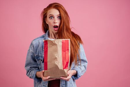 frowning surprised and amazesd long red-haired ginger woman holding paper eco bag package with chips...
