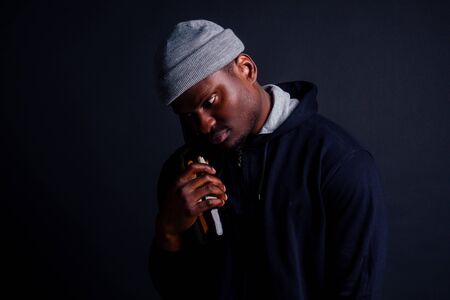 african american gue wearing gray hat and hoody hjlding botle with wine in srudio black background.alcoholism homeless bum conceptの写真素材