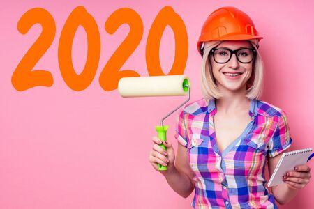 happy blonde woman in construction helmet hardhat painted wall with roller tool drawing with paint corange color . concept of New Yearの写真素材