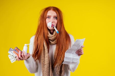 unwell redhaired ginger woman runny nose wearing knitted sweater and scarf with many pills in hand in studio yellow backgroundの写真素材