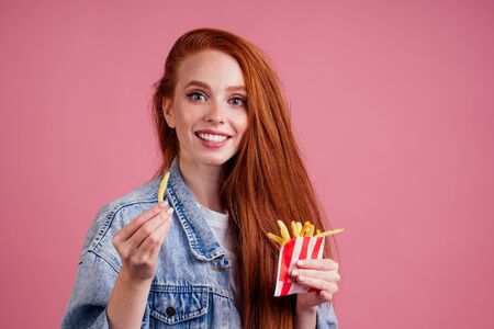 cute red long foxy haired ginger woman holding packaging cardboard box with french fries in pink studioの写真素材