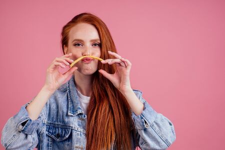 beautiful redhead ginger woman having fun with fry slice potato making moustache in studio pink...