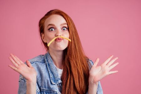 beautiful redhead ginger woman having fun with fry slice potato making moustache in studio pink...