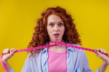 strangle redhaired ginger curly woman holding measure tape in studio yellow background.strangling her neckの写真素材