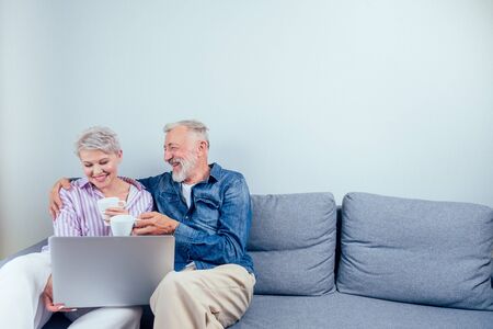elderly couple in love sitting gray couch in living room look at screen laptop and drinking cup of tea or coffee copyspaseの写真素材