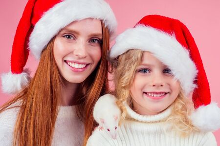 redhaired ginger woman with small little blond girl wearing santas hat holding many gift boxes with whire rat in studio pink backgroundの写真素材