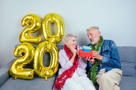 Happy beautiful elderly couple wearing christmas tinsel on neck celebrating new year at home in living roomの写真素材