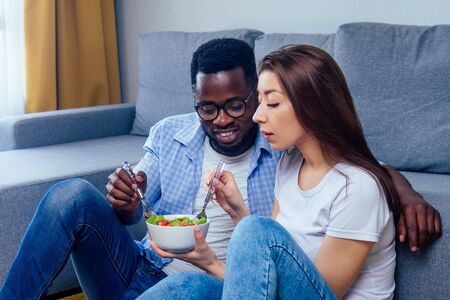 african american man eating salad with woman in living roomの写真素材