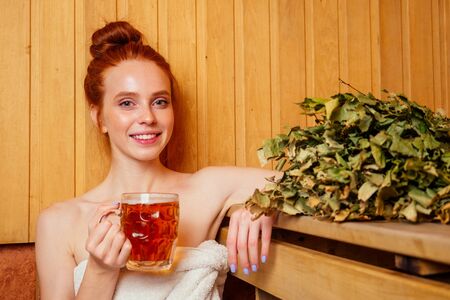 redhaired ginger woman steaming in the bathhouse with russian bunch broom drinking beers.she enjoying the sauna finnish spaの写真素材