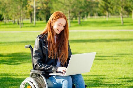 redhead ginger freelancer woman sitting in wheelchair and working by laptop in summer park