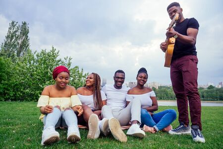 Group of smiling happy afro-american friends friends playing guitar outdoors picnic in the campaign