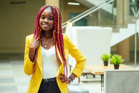 happy, afro american woman in a stylish yellow jacket and multi-colored dreadlocks pigtails workplace in a light large modern officeの写真素材