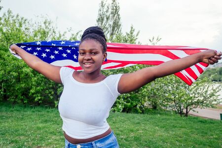 Smiling afro american woman holding USA flag and looking at camera autumn evening spring in the park by the lakeの写真素材