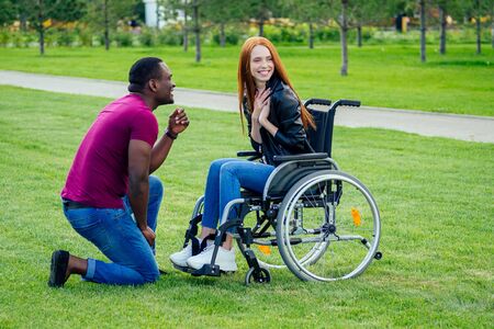 afro american man marriage proposal giving a ring to his redhaired ginger girlfriend.she sitting on wheel chair and surprised and amazedの写真素材