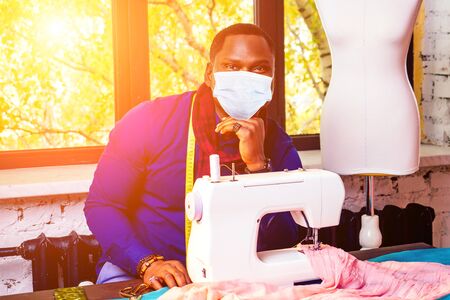 portrait of a handsome african man smiling seamstress with sewing machine. American man stylish designer working wear maskの写真素材