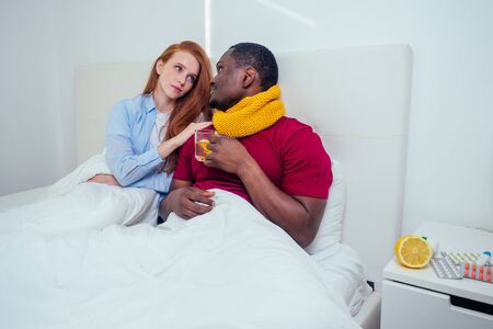Young couple in knitted scarf in bedroom, sick and measure temperatureの写真素材