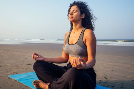 afro america woman with curly hair and closed eyes deep breathing and calming herself on empty morning beach after surya namaskarの写真素材