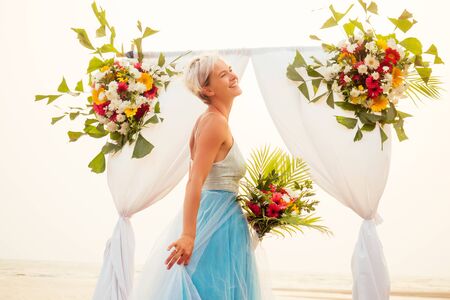 beautuful blonde short haired woman posing in blue tulle long train dress near wedding flower arch with tropical bouquet on sandy beachの写真素材