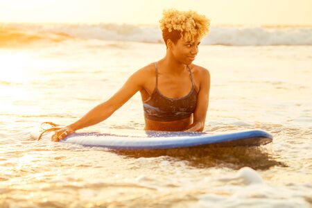 latin hispanic woman with blonde afro curls holdian a surfboard on beach.の写真素材