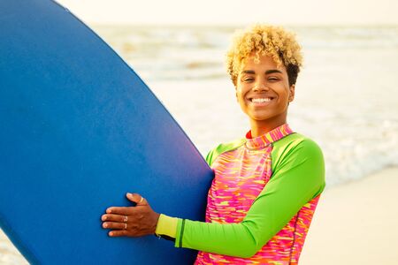 latin hispanic woman with blonde afro curls holdian a surfboard on beach