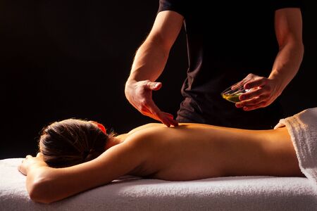 young indian woman lying on the table and getting ayurvedic massage with organic oil or honeyed in dark room.massagist male pouring out client backの写真素材