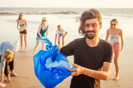 handsome indian man in the foreground collecting plastic from sand into the bag , his mixed race friends picking waste on background in Goa India beachの写真素材