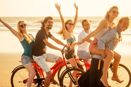 happy friends contest on cycle and sporty man giving their girlfriend piggyback rides summer freedom holiday on seaの写真素材