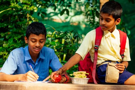 indian school boy writing on notepad doing homework , looking concentration . His friend with backpack sits on table and eating an apple in parkの写真素材