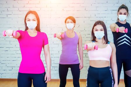 a group of four beautiful and sportive women in medical mask warming up and posing against a white brick wall in the gym. the concept of teamwork and fitnessの写真素材