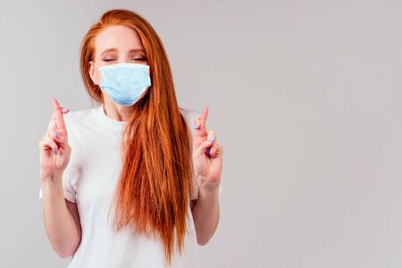 fingers crossed wishing the best in studio gray background redhair ginger woman feeling unhappy and wearing medical maskの写真素材