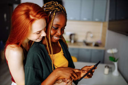 Lesbian couple looking at mobile phone and smiling in living room kitchenの写真素材