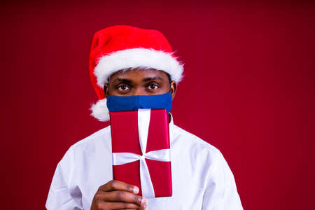 african american man in santas hat wearing mask in red studio backgroundの写真素材