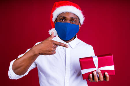 african american man in santas hat wearing mask in red studio backgroundの写真素材