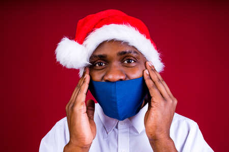 african american man in santas hat wearing mask in red studio backgroundの写真素材