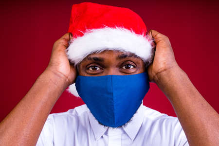 african american man in santas hat wearing mask in red studio background